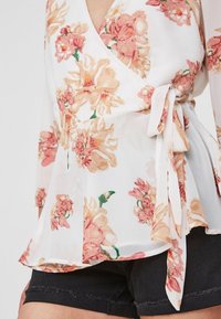 Blütenumhang-Oberteil aus leichtem, durchsichtigem weißen Stoff mit rosa und beigen Blumenmustern, ausgestattet mit einem Bindeband in der Taille und schwingenden Ärmeln.