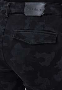 Los pantalones de mezclilla de camuflaje negro presentan una tela texturizada con un patrón sutil. Incluyen una etiqueta de cuero negro con "ALLSAINTS" grabada en la cintura.