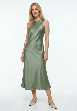 Vrouw met blond haar, gekleed in een mouwloze olivegroene satijnen midi-jurk en beige lage hakken, staand tegen een witte achtergrond.