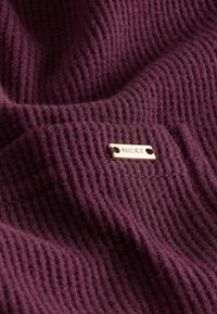 Dark purple ribbed fabric with a soft texture, featuring a small gold metal tag labeled "NEXT."
