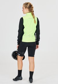 Atletische outfit met een felgroene, ritsjack met zwarte mouwen en zwarte fietsshorts met een hartslagpatroon. Zwarte helm in de hand gehouden.
