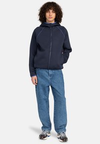 Navy zip-up hoodie with contrasting blue stitching and a hood, paired with loose, light blue jeans and patterned sneakers.