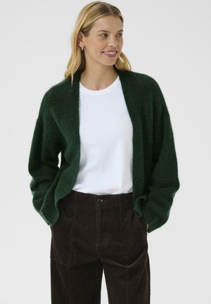 Grüner, flauschiger Cardigan mit offener Vorderseite, kombiniert mit einem weißen T-Shirt und braunen Cordhosen. Der Stoff wirkt weich und strukturiert.