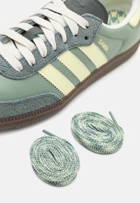 Green leather sneaker with grey suede accents, yellow striped design, and textured sole. Includes two pairs of patterned shoelaces.