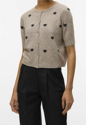 Cardigan beige à manches courtes boutonné avec petits motifs de cœurs noirs, porté avec un pantalon noir plissé taille haute.