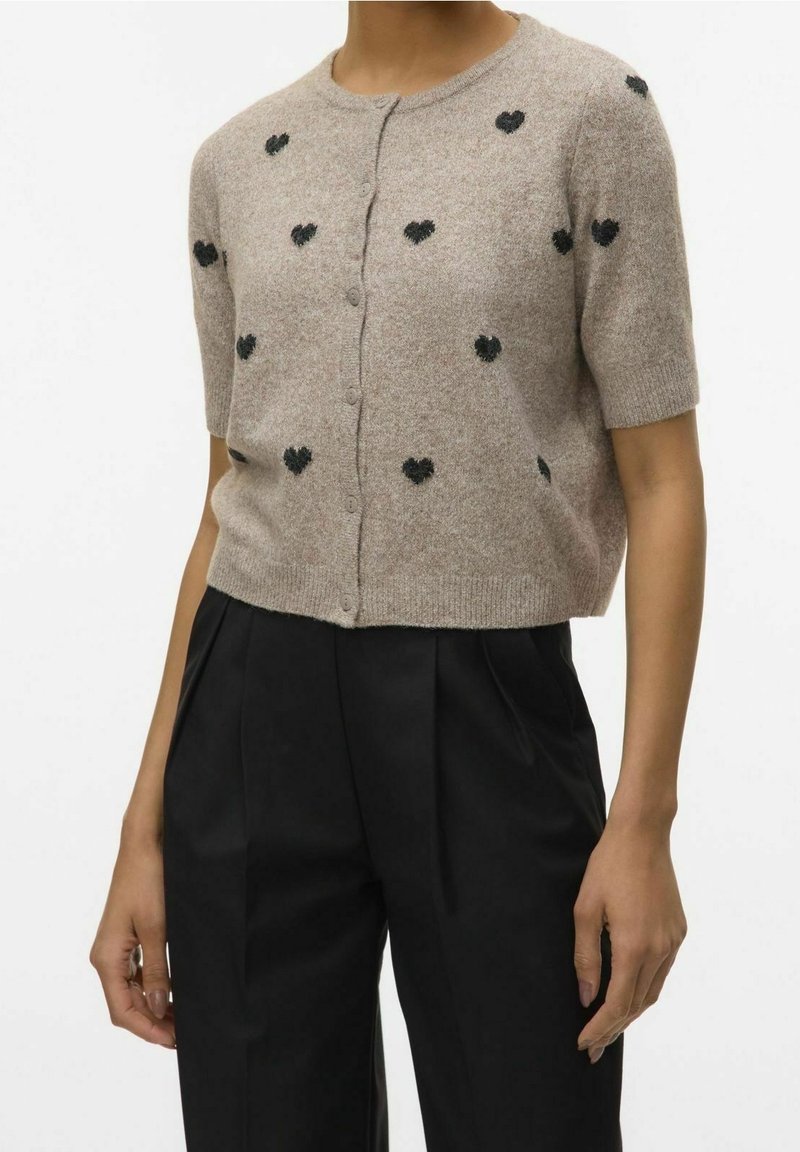 Cardigan beige à manches courtes boutonné avec petits motifs de cœurs noirs, porté avec un pantalon noir plissé taille haute.