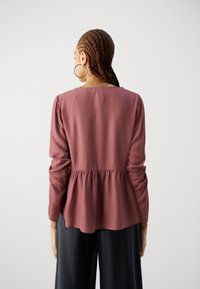 Top a maniche lunghe, di stile peplum, in un colore mauve tenue, realizzato in tessuto morbido con scollatura rotonda e dettaglio della vita arricciato.