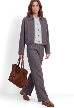 Frau geht, trägt eine graue Jacke und Hose mit einem weißen, geknöpften Oberteil, trägt eine große braune Wildlederhandtasche und braune Loafers.