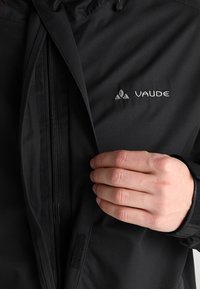 Veste Vaude noire avec fermeture éclair frontale, capuche ajustable et logo brodé. Le matériau semble texturé, durable et résistant aux intempéries.