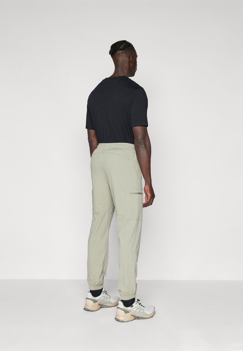 Pantaloni cargo verde chiaro con polsini elastici, tasche laterali e cintura con cordino, abbinati a una t-shirt nera a maniche corte e sneakers.