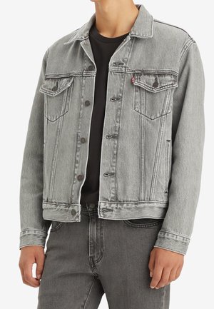 Grijze denimjas met klassieke kraag, voorzakken en metalen knopen. Licht vervaagde textuur, gecombineerd met een zwart shirt daaronder.