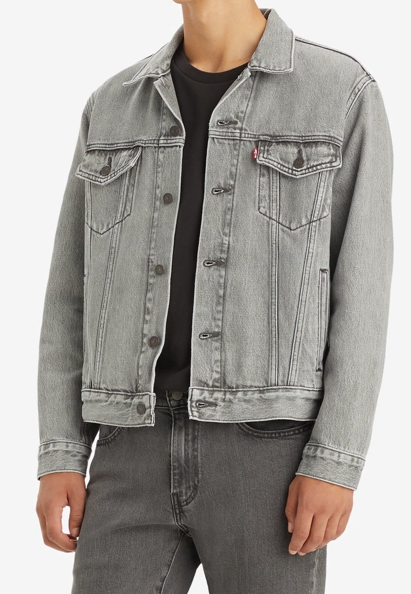 Giacca in denim grigia con colletto classico, tasche frontali e bottoni metallici. Tessuto leggermente sbiadito, abbinato a una camicia nera sotto.