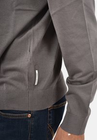 Suéter gris de punto con puños y dobladillo acanalados, presenta una abertura lateral y una pequeña etiqueta de marca en la costura del lado izquierdo. Combinado con jeans oscuros.