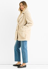 Beige faux fur coat with a wide collar, two front pockets, and button closure, paired with blue straight-leg jeans and black flats.