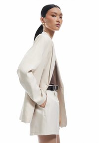 Beige blazer med oversized ærmer, parret med matchende shorts. Brun top underneath, sort bælte med guldbuckle. Glat stofstruktur.