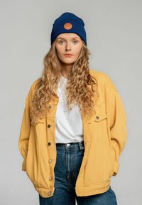 Żółta, oversize'owa kurtka z zatrzaskami, biała koszulka, niebieska dzianinowa czapka beanie z brązową naszywką, oraz niebieskie jeansy z wysokim stanem.