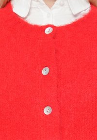 Suéter vermelho peludo com decote redondo. Apresenta botões prateados e uma superfície texturizada. Camisa branca com colarinho visível por baixo.