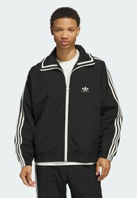 Schwarze Zip-Jacke mit weißen Streifen an den Ärmeln und am Kragen. Sie hat eine entspannte Passform, elastische Bündchen und ein gesticktes Logo auf der Brust.