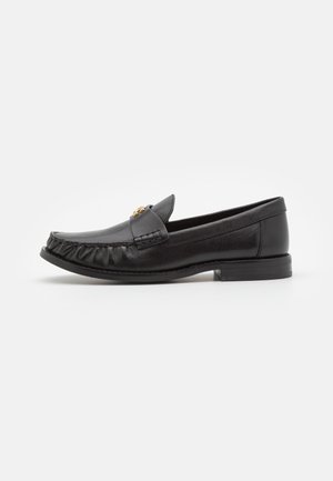 Mocasín de cuero negro con costuras tipo moc toe y un pequeño detalle metálico dorado en la parte superior, tacón plano y suela lisa sobre un fondo blanco.