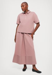 Helles rosa Poloshirt mit kurzen Ärmeln und einem Kragen, kombiniert mit einem passenden langen Rock. Der Stoff ist glatt und leicht strukturiert. Schwarze Ballerinas.