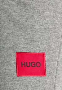 Tissu gris avec un tricot texturé, comportant un patch rectangulaire rouge avec l'inscription noire "HUGO" affichée de manière proéminente.