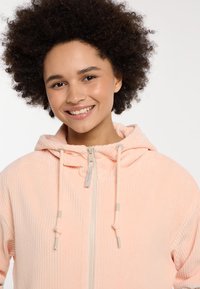 Lichtroze corduroy ritsjack met een trekkoordcapuchon, geribbeld textuur en een contrasterende ritsdetail.