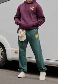 Mörkröd hoodie med gul logotyp, matchad med gröna sweatpants med samma logotyp. Krämfärgad keps placerad på midjan; vita sneakers.