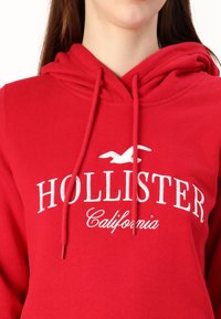 Sudadera roja de algodón, con un logo blanco que dice "HOLLISTER California," con capucha ajustable y puños acanalados.