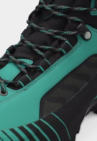 Κοντινή λήψη ενός μαύρου και τιρκουάζ ορειβατικού μποτακιού με κορδόνια με σχέδιο, υφή στη σόλα και ενισχυμένες οπές για τα κορδόνια.