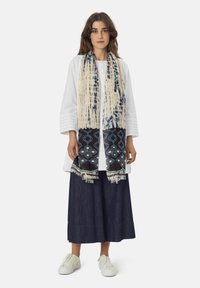 Scarf featuring a beige and navy pattern with geometric designs and fringed edges, worn over a white top and dark skirt.
