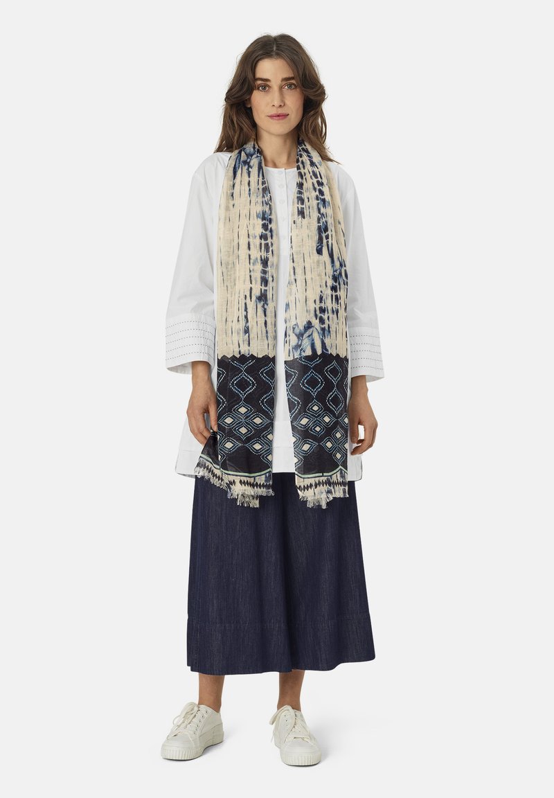 Écharpe présentant un motif beige et marine avec des designs géométriques et des bords frangés, portée sur un haut blanc et une jupe sombre.