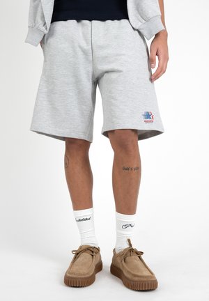 Mann trägt hellgraue knielange Shorts mit einem Logo am linken Bein, weiße Socken mit Text und beige Schnürschuhe.