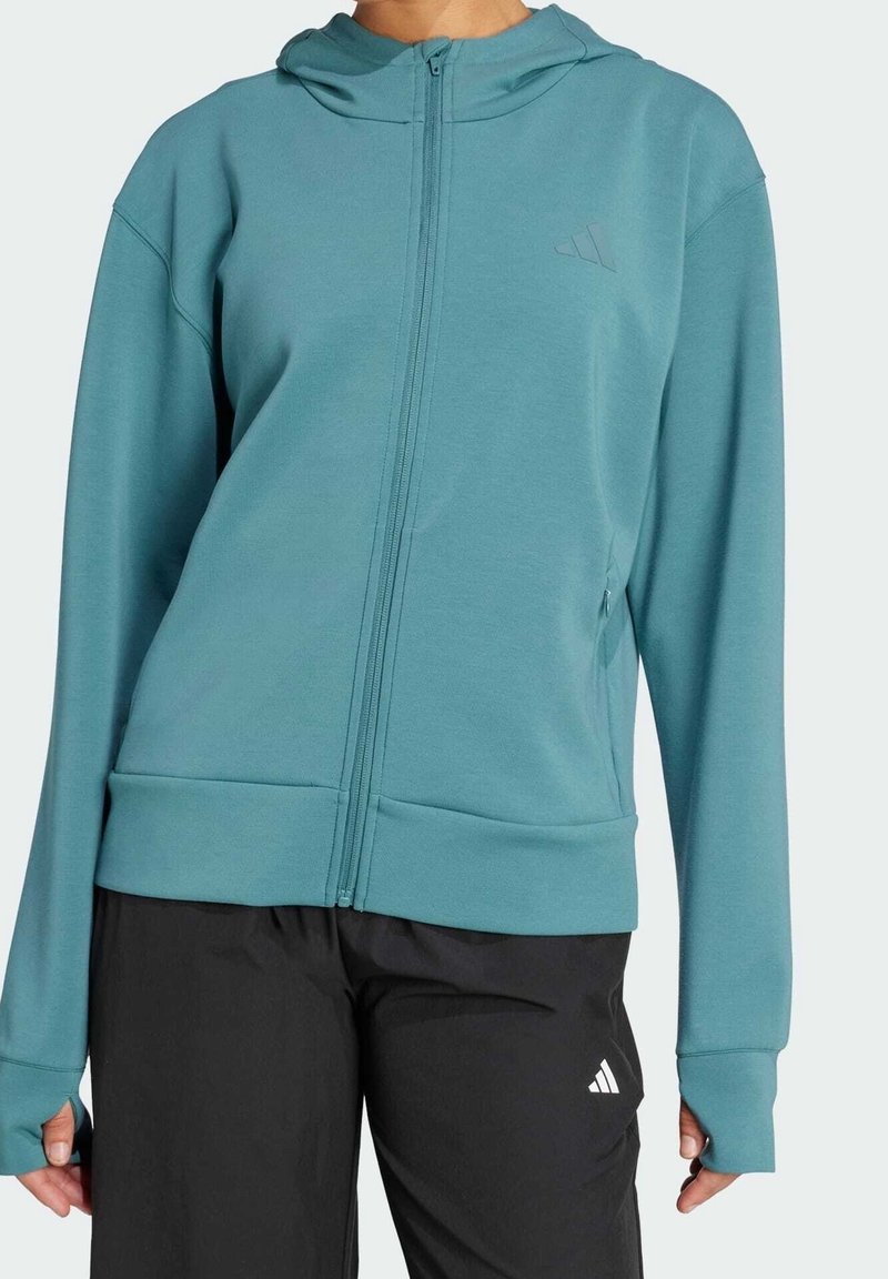 Sweat à capuche zippé bleu sarcelle avec trous pour les pouces, porté avec un pantalon de sport noir affichant un logo blanc sur la cuisse gauche.