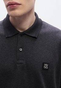 Σκούρο γκρι πόλο πουκάμισο κατασκευασμένο από υφασμάτινη υφή, με γιακά με κουμπιά, δ ύο κουμπιά και τετράγωνο ετικέτα με το λογότυπο "HUGO" στο στήθος.