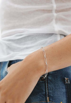 Pulsera de eslabones rectangulares plateados llevada en la muñeca izquierda sobre piel clara, combinada con una camiseta blanca y vaqueros azules de mezclilla.