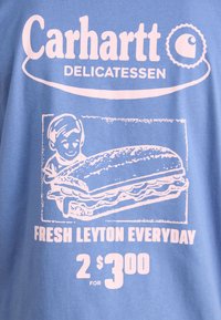 Šviesiai mėlynas medvilninis marškinėliai su rožiniu spauda grafika. Yra užrašas "Carhartt Delicatessen," vaikas su širdelėmis ir sumuštinis. Tekste yra nurodyta kaina.