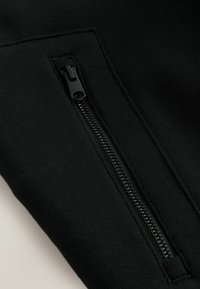 Schwarzer Sweatshirt-Stoff mit einer seitlichen Reißverschlusstasche. Der Reißverschluss ist schwarz mit einem strukturierten Zipper, der nahtlos in das Material integriert ist.