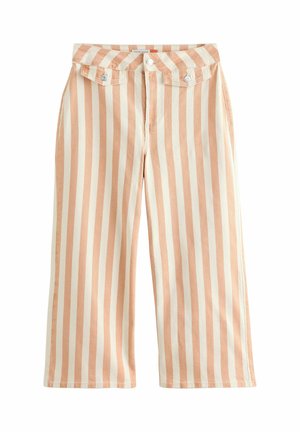 Kvinde iført hvid ærmeløs blondebluse, beige og hvide lodrette stribede vidde bukser og brune sandaler, stående med den ene arm hævet.