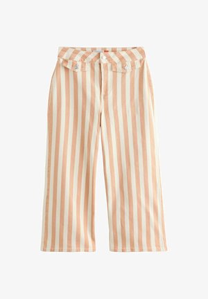 Frau trägt ärmelloses weißes Spitzenoberteil, beige-weiß gestreifte weit geschnittene Hose und braune Sandalen, steht mit einem erhobenen Arm.