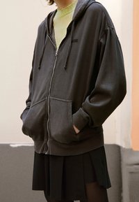 Gray zip-up hoodie with dual front pockets, drawstring hood, and ribbed cuffs, layered over a sheer green top and paired with a black skirt.