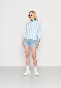 Camisa de manga longa azul clara e branca com riscas, calções de ganga claros e botas brancas até ao tornozelo. A modelo usa óculos de sol e um colar verde.