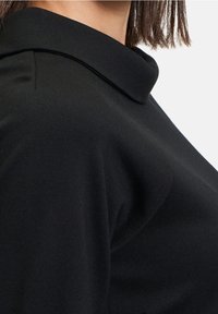 Černý top s tvarovaným límcem, vyrobený z hladké látky s texturovaným povrchem. Detailní pohled zdůrazňuje design límce a výstřihu.
