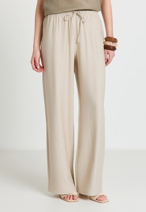Femme portant un pantalon ample beige avec cordon de serrage, un haut marron clair, des sandales beiges à bout ouvert et des bracelets en bois empilés au poignet droit.