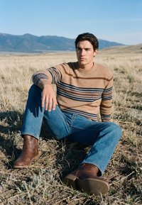Un jeune homme est assis sur de l'herbe sèche, portant un pull marron à rayures, un jean bleu et des bottes marron, avec des montagnes en arrière-plan sous un ciel clair.