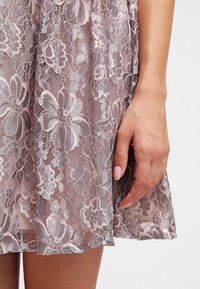 Robe en dentelle rose pâle avec des motifs floraux complexes. Présente une texture délicate, un ourlet court et une silhouette cintrée.