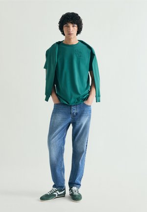 Joven de pie con las manos en los bolsillos, lleva una sudadera verde colgada sobre los hombros, camiseta verde, vaqueros azules y zapatillas deportivas verdes.