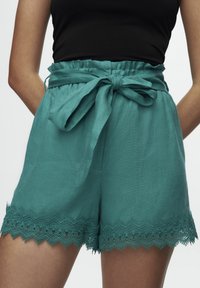 Pantalones cortos de color azul verdoso, hechos de un tejido ligero, con una cintura alta fruncida, lazo atado en el frente y dobladillo con encaje.