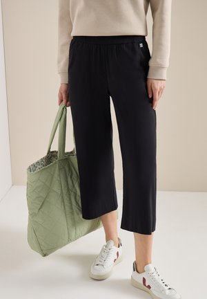 Persona con pantaloni neri corti, sneakers bianche con dettagli rossi, maglione beige, che tiene una grande borsa tote trapuntata verde chiaro.