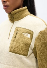 Φλις μπλούζα σε εκρού και ελιά, με ψηλό γιακά, τσέπη στο στήθος με φερμουάρ και κεντημένο λογότυπο στην τσέπη. Μαλακή υφή.