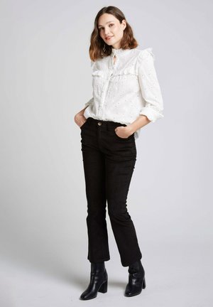 Blouse blanche brodée avec un col volanté, assortie à un pantalon en velours côtelé noir et des bottes à talons noires. Le modèle se tient les mains dans les poches.